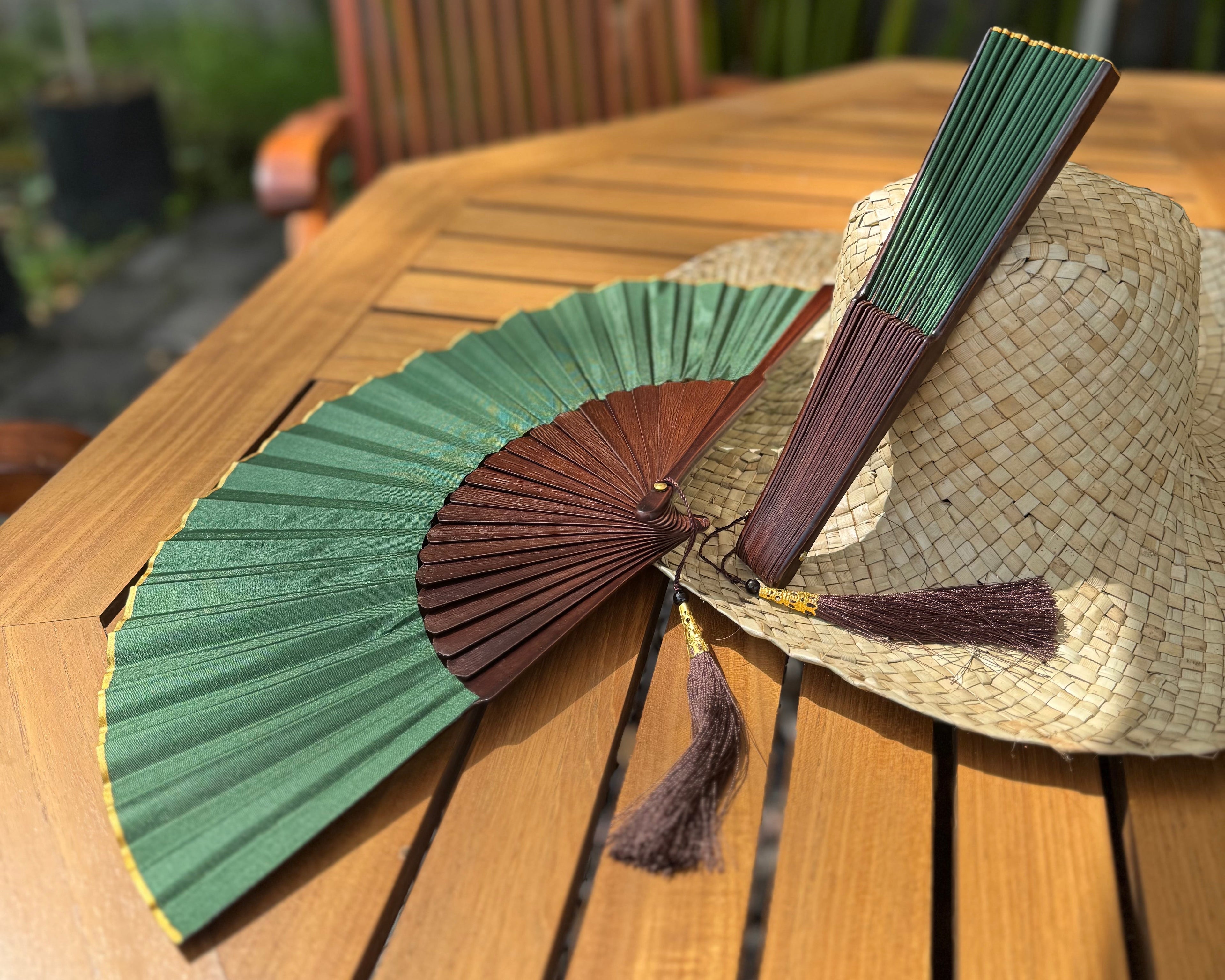 A traditional folding hand fan with green and dark brown ribs, resting on a wooden table next to a woven straw hat. The background shows a wooden chair and green foliage in a garden setting.